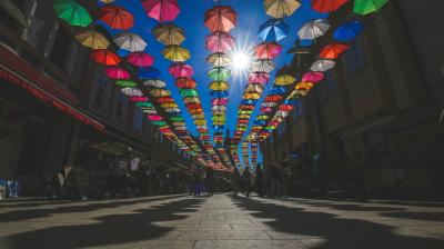 Colorful umbrellas are suspended above a pedestrian street, casting shadows on the ground; sunlight shines through the umbrellas and buildings line both sides of the street.