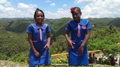 Two girls in blue school uniforms stand outdoors on a hilltop with green hills in the background. Subtitle reads, "If thunder roars stay indoors," in yellow text.