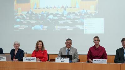 Six people sit at a conference table with nameplates, facing the camera, with a large screen showing an audience and meeting behind them.