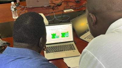 Two people look at weather or map data displayed on a laptop screen in an office meeting setting, with laptops and papers on the table.