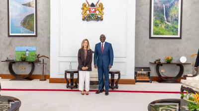 Two people stand side by side in a formal room with framed photographs, decorative furniture, and a national emblem on the wall behind them.