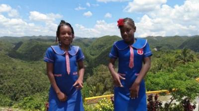 Two girls in blue school uniforms stand outdoors with green hills and a partly cloudy sky in the background.
