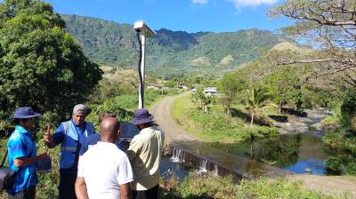Four people stand near a small river and road, next to a tall monitoring device, with mountains and trees in the background on a sunny day.