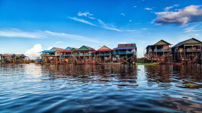 Colorful wooden houses on stilts line the edge of a calm body of water under a blue sky with scattered clouds.