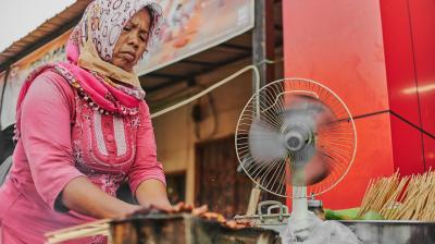 A woman in a pink dress and headscarf grills skewered food beside a spinning fan at an outdoor food stall.