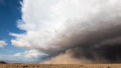 A large dust storm approaches across a dry, flat desert landscape under a partly cloudy sky.