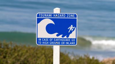 Blue and white sign reads "Tsunami Hazard Zone" and instructs to go to high ground or inland in case of earthquake, with wave and person graphic. Ocean is visible in the background.