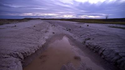 A muddy, shallow stream flows through a dry, cracked landscape under a cloudy sky at dusk, with minimal vegetation and a lone tree in the distance.