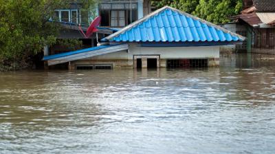 A house with a blue roof is partially submerged in floodwater, with only the roof and upper windows visible above the waterline.
