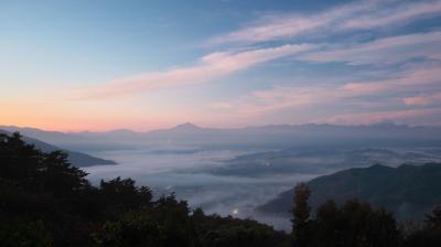 A landscape view of a valley covered in morning mist, with distant mountains under a colorful sunrise sky and scattered lights visible below.