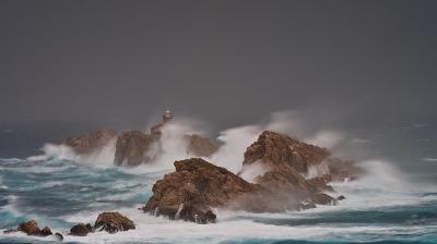 Waves crash against rocky outcrops in a stormy sea, with a lighthouse visible on one of the rocks under a dark, cloudy sky.