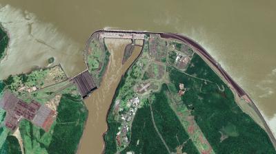Satellite view of a large dam spanning a river with adjacent buildings, green vegetation, and brown water flowing through the structure.