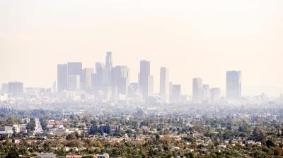 Downtown city skyline with tall buildings partially obscured by smog, seen from a distance above residential and commercial areas.