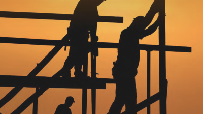 Three construction workers are silhouetted against an orange sunset sky as they work on a building framework.