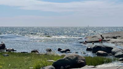 Rocky shoreline with grass in the foreground, waves in the sea, a few people near the water, and a sailboat visible in the distance under a partly cloudy sky.