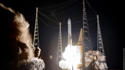 A rocket launches at night, producing bright flames and smoke, surrounded by tall support towers against a dark sky.