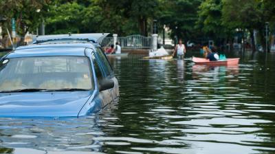 A blue car is partially submerged in floodwater, while people use boats to navigate a flooded street lined with trees in the background.