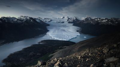 A large glacier flows between mountain ranges, ending in a lake under a cloudy sky, with rocky terrain in the foreground.