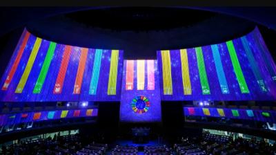 The interior of a large auditorium displays multicolored vertical light panels and a circular emblem on the wall above seated attendees.