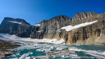 Rocky cliffs rise above a partially frozen turquoise lake with floating ice and patches of snow under a clear blue sky.