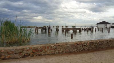 A calm lake scene under a cloudy sky, showing partially submerged stone pillars and vegetation near the shore. A low stone wall runs along the sandy foreground, and a small white building with a dark roof stands on stilts over the water in the distance.