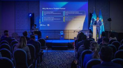 A speaker presents information on a screen titled "Why We Are a Trusted Partner" to an audience seated in a conference room with blue lighting.