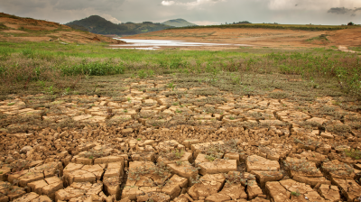 Dry, cracked earth in the foreground with sparse vegetation, a receding body of water in the middle ground, and hills under a cloudy sky in the background.