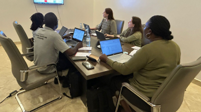 Five people sitting around a conference table with laptops, viewing a large screen displaying a spreadsheet in a meeting room.