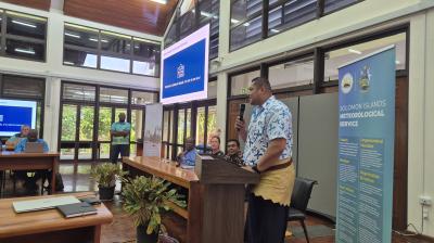 A man speaks at a podium during an indoor event, with attendees seated at tables and banners displaying "Solomon Islands Meteorological Service" visible in the background.
