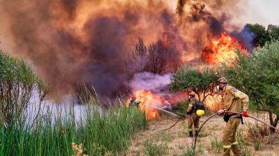 Two firefighters in protective gear use hoses to battle a large wildfire, with thick smoke and tall flames consuming vegetation around them.