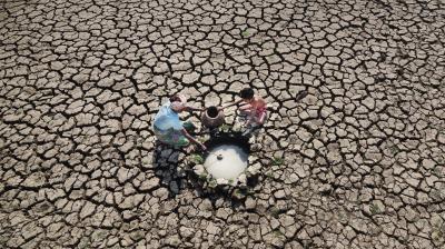 Three people collect water from a small well in the middle of a cracked, dry landscape, indicating drought conditions.