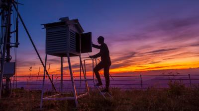 A person checks equipment inside a weather station at sunset, standing on a small ladder beside a fenced area and tall antenna structure.