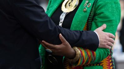 Two people in formal attire embrace; one wears a vibrant green jacket with patterned sleeves and large jewelry, while the other's suit is dark and plain.