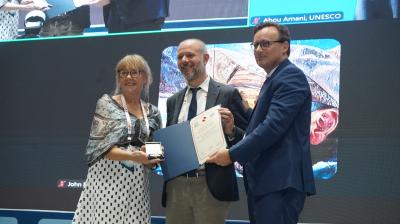 Three people stand on stage holding certificates and an award, with a large screen displaying their image in the background.