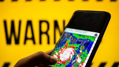 A hand holds a smartphone displaying a weather radar map in front of a yellow background with the word "WARNING" in bold black letters.