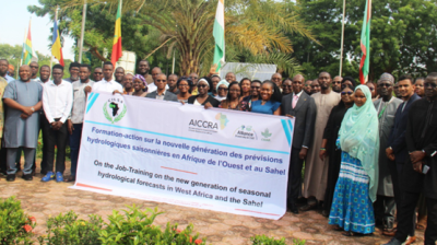 A large group of people pose outdoors, holding a banner about hydrological forecast training in West Africa and the Sahel, with flags and trees in the background.