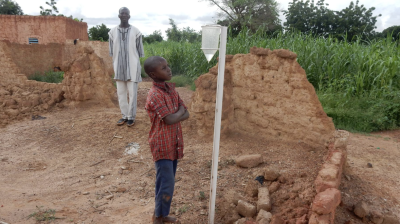 A boy observes a rain gauge in a rural area with an adult standing behind him; damaged mud-brick structures and green crops are visible in the background.