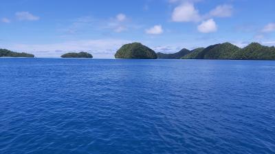 A calm, bright blue ocean with several small, green, forested islands under a partly cloudy sky.