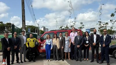 A group of people in business attire stand in a line outdoors in front of a red vehicle, with trees and a partly cloudy sky in the background.