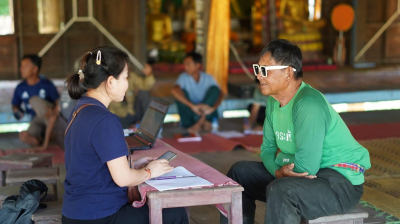 A woman interviews an older man in sunglasses indoors; she holds a phone and notebook, while others sit in the background.