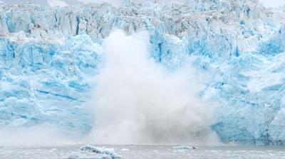 Large chunk of glacier ice collapsing into the water, creating a splash and mist at the base, with blue and white ice formations in the background.