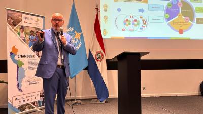 A man in a blue suit speaks into a microphone next to presentation slides, standing by the UN and Paraguayan flags at a conference.