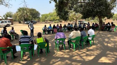 A group of people sit in a large circle on green plastic chairs outdoors, while one person stands and speaks; trees and vehicles are visible in the background.