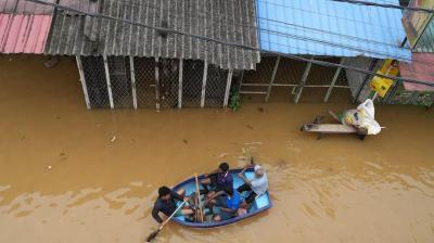 Three people row a small boat through floodwaters in front of submerged buildings, while a man sits on a table with bags nearby.