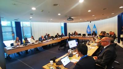 A group of people sit around a large conference table with laptops and documents in a meeting room, featuring United Nations flags in the background.