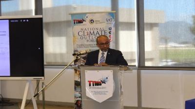 A man in a suit stands at a podium speaking during the National Climate Outlook Forum, with a presentation screen and banner in the background.