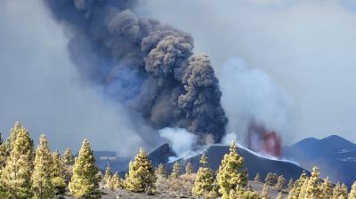 A volcano erupts, emitting thick black smoke and ash into the sky, with some trees visible in the foreground on a rocky landscape.