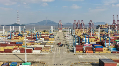 Aerial view of a large shipping port with stacked cargo containers, cranes, trucks, and distant water and hills under a partly cloudy sky.