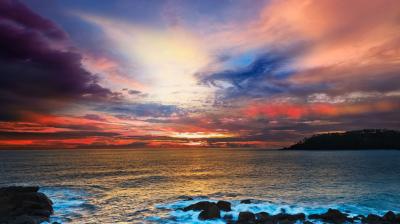 Colorful sunset over the ocean with waves crashing against rocks in the foreground and partly cloudy sky filled with vibrant hues of orange, pink, blue, and purple.