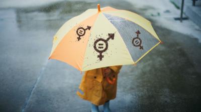 A child in a yellow raincoat stands under a yellow and orange umbrella with gender symbol designs on a rainy day.